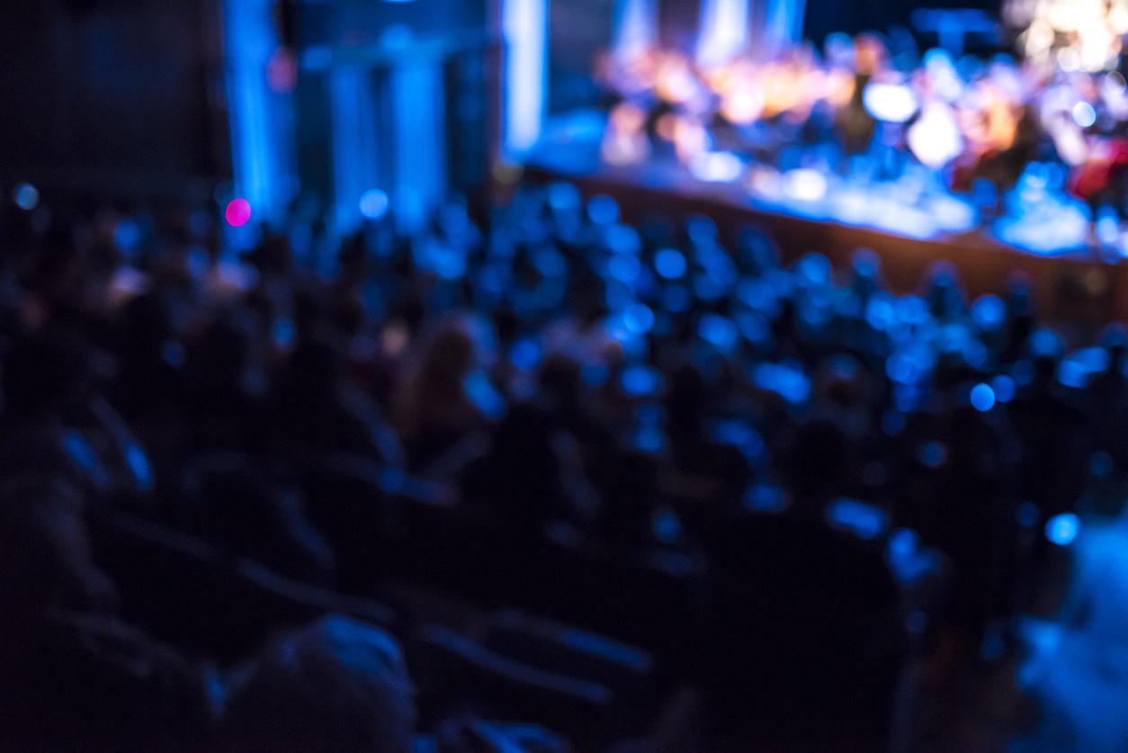 Defocused shot of an audience listening to a concert indoors. Orchestra is on the stage. Blue lights are  shining.