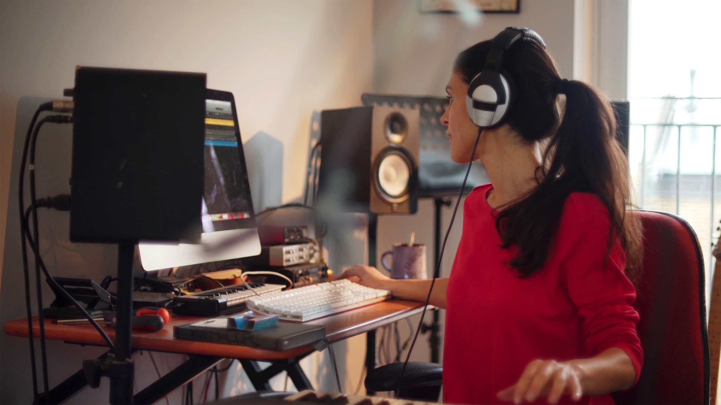 A good looking Hispanic woman is composing music in her private home studio. She is using a computer &amp; music keyboard to compose and is surrounded by musicians’ equipment.