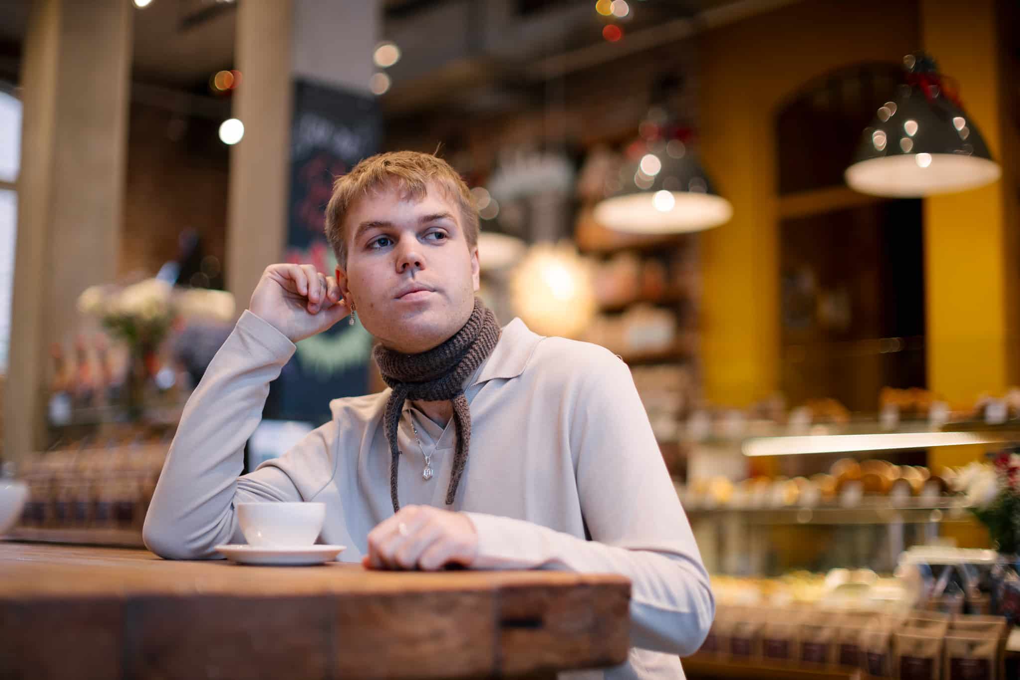 En mann i beige poloskjorte og skjerf sitter med en kaffekopp i en kafé.