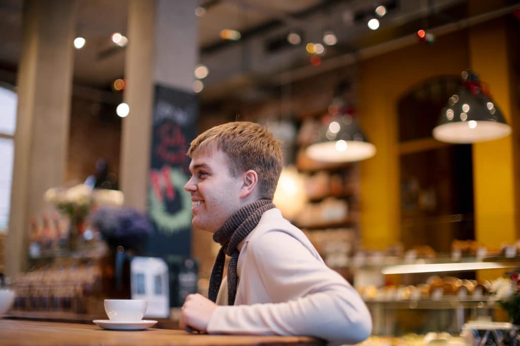 En mann i beige poloskjorte og skjerf sitter med en kaffekopp i en kafé. Han ser bort mot noen og ler. 