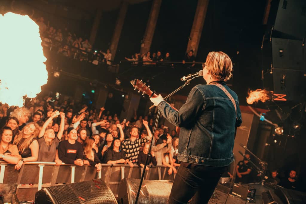 TONO_sommerlaater_VidarVilla_2.jpg – En blond mann med jeansjakke spiller gitar og synger foran et stort publikum.